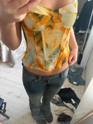 Korsettopp från PrettyLittleThing - Säljer en snygg korsettopp från PrettyLittleThing med blommigt mönster i orange, gul, grön och vitt. 