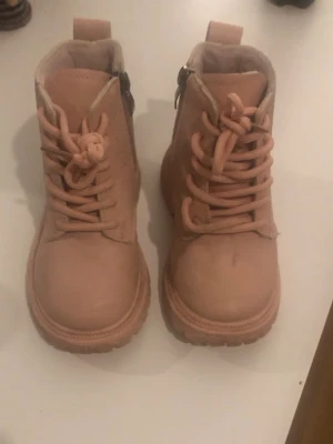 Rosa boots från Cat& Jack,med snörning och dragkedja - Säljer ett par ljusrosa boots med grov sula, snörning framtill och dragkedja på sidan för enkel på- och avtagning. Skorna är tillverkade i syntetmaterial med rund tå och platt sula. Insidan är fodrad för extra komfort och värme.
