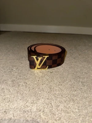 Brunt rutigt bälte med guldfärgat spänne - Elegant och tidlöst bälte i klassiskt brunt rutmönster med ett stilrent guldfärgat spänne som verkligen lyfter hela outfiten. Perfekt accessoar för dig som vill addera en lyxig känsla till både jeans och mer dressade looks.  Bältet är i fint skick och redo att användas direkt. En riktig statement-detalj som aldrig går ur stil.