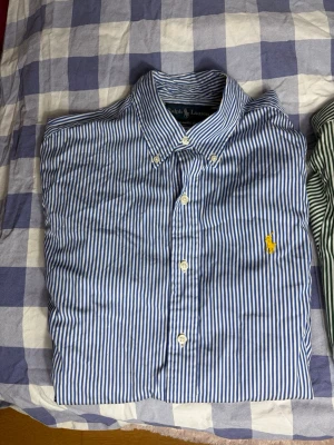 Blåvit randig skjorta från Ralph Lauren - Snygg blåvit randig skjorta från Ralph Lauren i klassisk passform. Skjortan har knappar framtill, button-down krage och den ikoniska gula Polo-loggan broderad på bröstet. Perfekt för dig som gillar stilrena och tidlösa plagg.