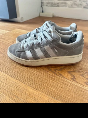 Grå Adidas Campus sneakers mocka - Säljer ett par grå Adidas Campus sneakers i mocka med klassiska vita ränder på sidorna och Adidas-logga bak. Skorna har rund tå, platt sula och breda vita snören. Perfekta för dig som gillar retrovibbar och streetstyle. Obs. Skorna har  hål inuti 