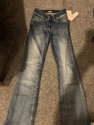 Blå bootcut jeans från Nelly, stl 34 - Säljer ett par blå bootcut jeans från Nelly i storlek 34. Modellen är lågmidjad med snygga rosa broderade detaljer på bakfickorna och dubbla knappar fram. Jeansen har en klassisk tvättad look och är gjorda i mjukt denimtyg.