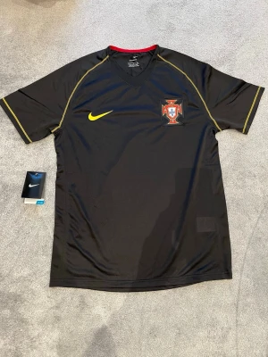Svarta Nike fotbollströja Portugal S - Säljer en svart Portugal fotbollströja från Nike med gul logga och broderat landslagsemblem på bröstet. Tröjan har gula sömmar, röd insida på kragen och är tillverkad i Sphere Dry-polyester som andas. Perfekt för dig som älskar fotboll och vill ha en snygg landslagströja.