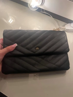 väska Anna field  - Snygg svart axelväska från Anna field med quiltad design och guldfärgad kedja som axelrem. Väskan har ett stilrent utseende med ett litet metallmärke framtill och mjukt material som känns lyxigt. Perfekt storlek för det viktigaste och passar till många olika outfits.