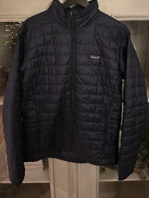 Mörkblå quiltad pufferjacka Patagonia - Quiltad pufferjacka från Patagonia i mörkblått med dragkedja framtill och två sidofickor med dragkedja. Jackan har hög krage och klassisk Patagonia-logga på bröstet. Perfekt för kyliga dagar och enkel att styla med hoodies eller t-shirts.