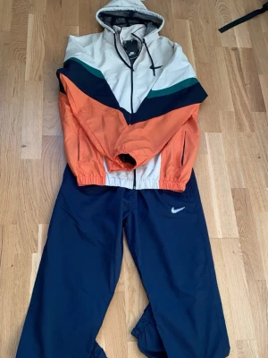 Nike träningsset med färgblock - Nike träningsset med jacka och byxor. Jackan har huva, dragkedja och färgblock i beige, orange, marinblå och grönt. Byxorna är marinblå med Nike-logga på låret. Perfekt för sport eller chill. Materialet är syntet och jackan har elastiska muddar.