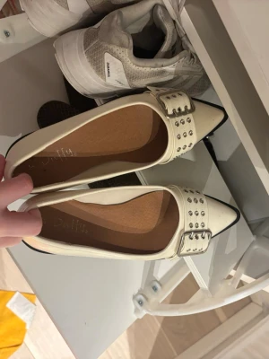 Vita ballerinaskor med nitar och spänne - Säljer ett par vita ballerinaskor från Duffy med spetsig tå och stora silvriga spännen dekorerade med nitar. 