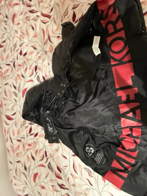 Svart pufferjacka från Michael Kors - Säljer en svart pufferjacka från Michael Kors med stor röd logga på insidan. Jackan har huva, dragkedja och två sidofickor. Materialet är glansigt och känns riktigt modernt. Perfekt för dig som vill ha en snygg och varm jacka med street vibe.
