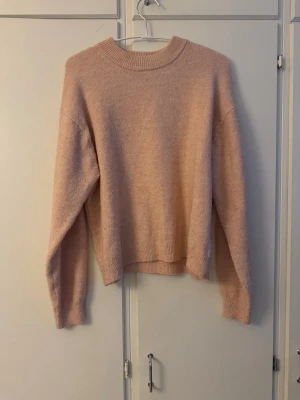 Ljusrosa stickad tröja med rund hals - Mysig ljusrosa stickad tröja med rundad hals och ribbade muddar vid ärmslut och nederkant. Tröjan har en loose passform och långa ärmar, perfekt för kyliga dagar. Enkel och stilren design som är lätt att matcha med olika outfits.