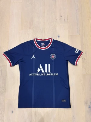 PSG x Jordan blå fotbollströja - Snygg blå PSG fotbollströja från Jordan med vita och röda detaljer på krage och ärmslut. Tröjan har Paris Saint-Germain-logga på bröstet, Jordan-logga och stor vit ACCOR LIVE LIMITLESS-tryck framtill. Perfekt för dig som älskar fotboll och streetwear.