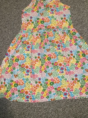 Blommig kortklänning från Amy & Lucy - Färgglad kortklänning från Amy & Lucy i storlek 122/128. Klänningen har ett blommigt mönster i rosa, blått, grönt, gult och orange på vit botten. Ärmlös modell med rund hals och mjukt bomullsmaterial som känns skönt mot huden. 