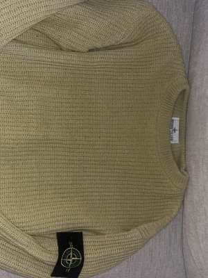 Beige stickad tröja från Stone Island - Säljer en beige stickad tröja från Stone Island med klassisk rund hals och den ikoniska logotypen på ärmen. Tröjan har grovstickad struktur och ribbade muddar vid halsen. Perfekt för dig som gillar streetwear och vill ha något snyggt och bekvämt.