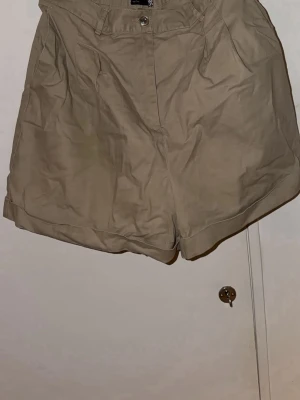 Beige höga shorts från ASOS Design - Säljer ett par beige höga shorts från ASOS Design, modell Tall. De har klassisk knapp och dragkedja framtill, fickor på sidorna, samt uppvikta benslut. Materialet är mjuk bomull med lite stretch för extra komfort.