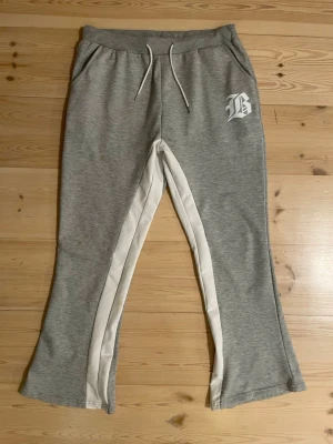 Grå denim bootcut mjukisbyxor med vit detalj - Snygga grå denim mjukisbyxor med bootcut och vita paneler längs benen. Byxorna har snörning i midjan och en vit tryckt bokstav på höger sida. Perfekta för chill dagar och har en skön passform med mjukt material.