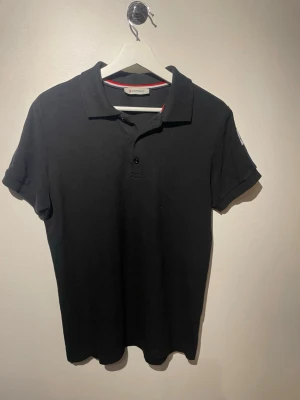 Svart Moncler polo piké  - Säljer en svart polotopp från Moncler i storlek XL. Toppen har klassisk krage, korta ärmar och två knappar framtill. På axlarna finns röda och vita ränder som ger en sportig vibe. Materialet är mjuk bomull och toppen har en normal passform. Riktigt liten i storlek