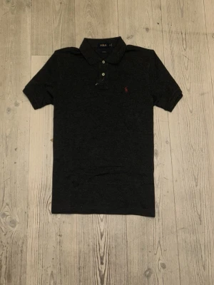 Mörkgrå pikétröja från Polo Ralph Lauren - Snygg mörkgrå pikétröja från Polo Ralph Lauren med klassisk krage, två knappar och röd broderad logga på bröstet. Tröjan är i bomull och har korta ärmar, ett bra grish plagg