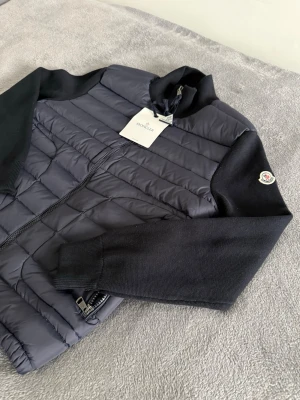 Moncler cardigan  - Snygg svart pufferjacka från Moncler med stickade ärmar och quiltad framsida. Jackan har dragkedja framtill, ståkrage och Moncler-logga på ärmen. Materialmix av polyester och stickad textil ger en modern vibe. Perfekt för dig som vill ha något stilrent och bekvämt.