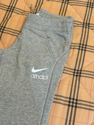 Grå mjukisbyxor från Nike Athletic Dept - Grå mjukisbyxor från Nike Athletic Dept med vit logga och text på vänster ben. Byxorna har snörning i midjan och två sidofickor. Tillverkade i mjukt bomullsmaterial som är skönt att ha på sig. Perfekta för chill eller träning.