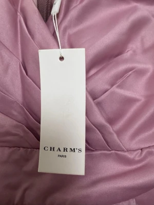 Ljusrosa långklänning från Charm's - Säljer en elegant ljusrosa långklänning från Charm's Paris. Klänningen har omlottliv med breda axelband och v-ringad hals, samt ett svepande, utsvängt kjolparti i glansigt satinmaterial. Perfekt för speciella tillfällen och ger en lyxig känsla.