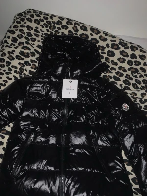 Svart pufferjacka från Moncler S - Svart glansig pufferjacka från Moncler med huva och dragkedja framtill. Jackan har två fickor med dragkedja och Moncler-logga på ärmen. Materialet är syntet och jackan har en quiltad design som ger en cool och modern look.