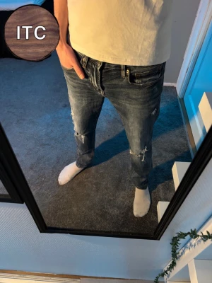 Jeans med slitningar  - Säljer ett par mörkblå skinny jeans från American Eagle med slitna detaljer och snygga rippade partier på benen. Jeansen har klassisk femficksdesign, låg midja och är tillverkade i mjukt bomullsmaterial med stretch för skön passform.