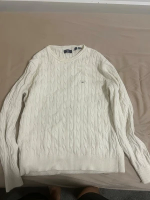 Vit kabelstickad tröja från GANT - Säljer en vit kabelstickad tröja från GANT med rund hals och diskret logga broderad på bröstet. Tröjan har långa ärmar och breda ribbade muddar vid nederkant och ärmslut. Perfekt för dig som gillar klassisk och stilren look.