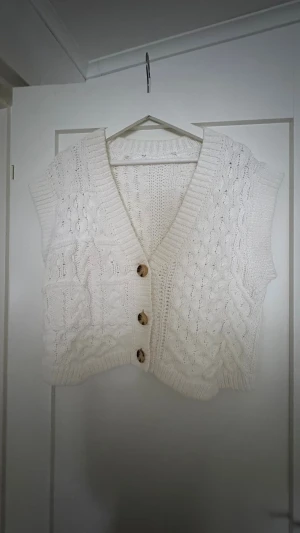 Vit stickad väst med knappar - Säljer en vit stickad väst med grovt kabelmönster och tre stora, mönstrade knappar framtill. Västen har v-ringad hals och ribbade kanter. Modellen är loose och croppad, perfekt att slänga över en t-shirt eller topp för en chill look.