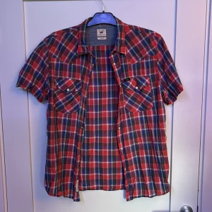 Rutig kortärmad skjorta från Lee - Snygg rutig skjorta från Lee i slim fit-modell. Skjortan är kortärmad och har röda, blå och vita rutor samt två bröstfickor med knappar. Perfekt för dig som gillar en klassisk men cool stil. 