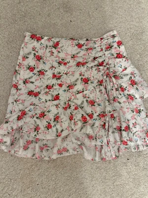 Vit blommig volangkjol med snörning - Säljer en kort kjol i vit med blommigt mönster i rosa och rött. Kjolen har volangdetaljer längs nederkanten och en snygg snörning på sidan som ger en extra touch. Perfekt för dig som gillar romantisk och feminin stil.
