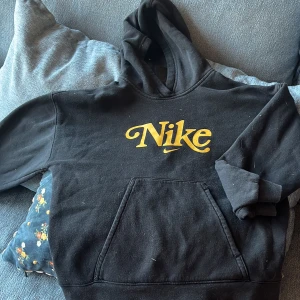 Svart Nike hoodie med gul logga - Svart hoodie från Nike med stor gul logga framtill. Tröjan har huva, känguruficka och en loose fit-passform. Perfekt för dig som gillar streetwear och vill ha en skön och stilren look.