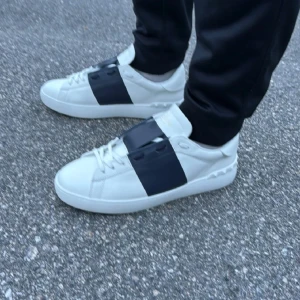 Valentino Garavani vita sneakers - Säljer ett par snygga Valentino Garavani sneakers i vitt med breda mörkblå paneler över snörningen. Skorna har rund tå, platt sula och coola nitar längs hälen. Tillverkade i skinn med klassisk snörning och stilren design.