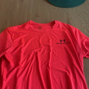 Orange Under Armour UA Rush t-shirt - Säljer en orange Under Armour UA Rush t-shirt i loose fit. T-shirten har korta ärmar, rund hals och är tillverkad i ett syntetmaterial som andas. Diskret logga på bröstet och i nacken. Perfekt för träning och sportiga aktiviteter.