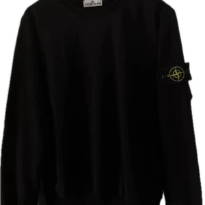 Svart sweatshirt från Stone Island - Svart sweatshirt från Stone Island med klassisk rund hals och ikonisk logga på vänster ärm. Tröjan är i bomull och har ribbade muddar vid ärmslut och nederkant. Perfekt för dig som gillar stilrena och trendiga plagg. 