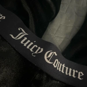 Svarta Juicy Couture mjukisbyxor S - Svarta mjukisbyxor från Juicy Couture i glansigt material med bred resår i midjan där Juicy Couture-loggan syns tydligt. Byxorna har en loose passform och är tillverkade i 95% polyester och 5% elastan för extra stretch och komfort.