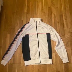 Vit och blå Kappa track jacket M - Snygg vit och blå tracksuit/ziptröja från Kappa i storlek medium. Jackan har hel dragkedja, höga muddar och klassisk Kappa-logga på bröstet och ryggen. Röd detaljlinje och mörkblåa paneler på sidorna samt ränder längs ärmarna som ger en sportig retro vibe.