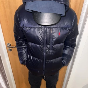 Mörkblå dunjacka från Polo Ralph Lauren - Snygg mörkblå dunjacka från Polo Ralph Lauren med huva och dragkedja. Jackan har en klassisk pufferstil, diskret logga i rött på bröstet och flera praktiska fickor. Perfekt för kalla dagar och riktigt skön att bära.