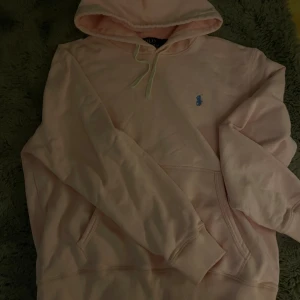 Ljusrosa hoodie från Polo Ralph Lauren - Säljer en ljusrosa hoodie från Polo Ralph Lauren med klassisk huva och snörning. Tröjan har en broderad blå logga på bröstet, känguruficka och ribbade muddar. Perfekt för dig som gillar stilrena och bekväma plagg.