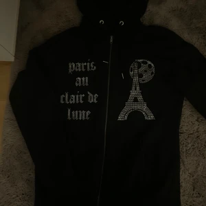 Svart hoodie med strass och tryck - Svart hoodie från BLINGLUCID med dragkedja, huva och snyggt strasstryck av Eiffeltornet och måne på framsidan. Texten 'paris au clair de lune' i glittriga bokstäver ger en unik vibe. Perfekt för dig som gillar statement-plagg.