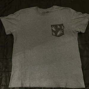 Grå t-shirt från Vans med ficka - Snygg grå t-shirt från Vans i bomull med korta ärmar och en svart bröstficka med grått blad-mönster och Vans-logga. Perfekt för en chill och avslappnad stil.