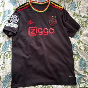 Ajax svart kortärmad fotbollströja - Ajax fotbollströja från Adidas i svart polyester med röda detaljer och Ziggo-logga på bröstet. Färgglada ränder i rött, gult och grönt vid krage och axlar. Champions League Foundation-märke på ärmen och Ajax-emblem med tre stjärnor. Perfekt för dig som gillar snygg design och fotboll.     ‼️‼️ Inte äkta ‼️‼️