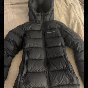 Svart dunjacka Peak Performance S - Säljer en svart dunjacka från Peak Performance i storlek S. Jackan har huva, dragkedja framtill och är quiltad för extra värme. Perfekt för kalla dagar och har en stilren look med diskret logga på bröstet. 3 fickor på utsidan och 1 på insidan. En ficka är krånglig att stänga