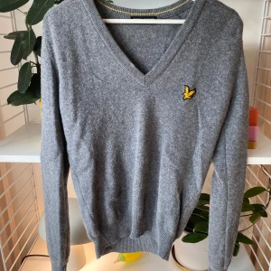 Grå v-ringad tröja från Lyle & Scott - Grå v-ringad tröja från Lyle & Scott med klassisk gul broderad fågel på bröstet. Tröjan är stickad och har långa ärmar samt ribbade muddar. Perfekt för lager-på-lager och snygg till både jeans och chinos.