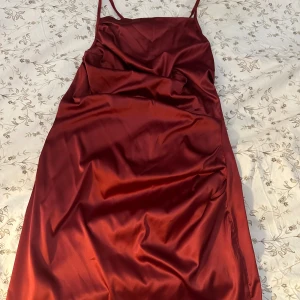 Vinröd satin slipklänning - Snygg vinröd slipklänning i satin med tunna axelband och rak passform. Klänningen har en slät, glansig yta och justerbara band i ryggen för perfekt passform. Perfekt för dig som vill ha en stilren och elegant look.