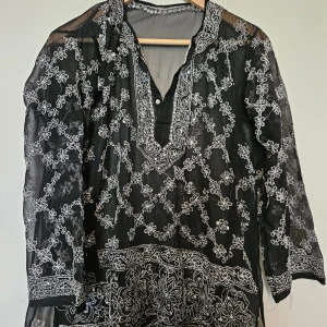 Svart blus med broderade blommor - Säljer en transparent svart blus med vita broderade blommor och dekorativa detaljer runt halsringningen. Blusen har v-ringning med knapp och trekvartsärmar. Perfekt för dig som gillar unika och luftiga plagg med snygga broderier.