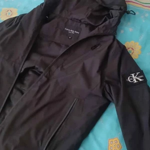 Svart vindjacka från Calvin Klein Jeans - Svart vindjacka från Calvin Klein Jeans med huva och dragkedja framtill. Jackan har logotyp på vänster ärm och praktiska fickor med dragkedja. Perfekt för blåsiga dagar och har en stilren, modern look.