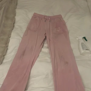 Rosa mjukisbyxor från Juicy Couture - Säljer ett par klassiska rosa mjukisbyxor från Juicy Couture med broderad JC-logga på fickan. Byxorna har snörning i midjan, stora fickor fram och bak med knappdetaljer. Mjukt och glansigt material som ger en lyxig känsla. Använd runt 10 gånger men i bra skick!