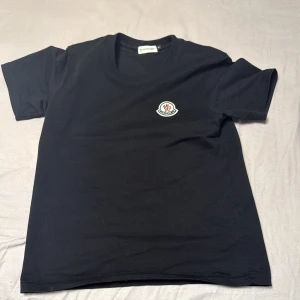 Svart Moncler t-shirt med logga - Svart t-shirt från Moncler i mjuk bomull med klassisk rund hals och korta ärmar. På bröstet sitter Monclers ikoniska broderade logga i vitt, blått och rött. Enkel och stilren design som passar perfekt till jeans eller shorts.