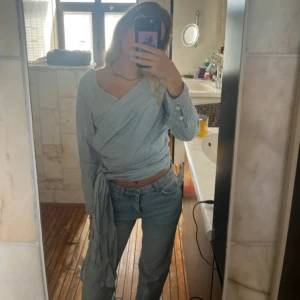 Randig omlottopp med knyt - Säljer en ljusblå och vit randig omlottopp med långa ärmar och snygg knytning i midjan. Toppen har v-ringad hals och smala ränder som ger en fräsch look. Perfekt att styla med jeans för en chill vibe.