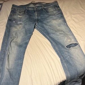 Replay Anbass ljusblå jeans, waist 38 - Säljer ett par Replay Anbass jeans i ljusblå tvätt med feta slitningar och hål på ena benet. Klassisk femficksmodell med snygga detaljer och läderpatch bak. Tillverkade i bomull och har en relaxed vibe. Perfekta för dig som gillar en lite mer slitiga jeans med väldigt snygga, passar perfekt med min vita stone island vindjacka som också ligger uppe på min plick😃