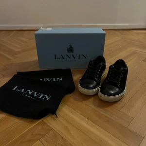 Svarta sneakers från Lanvin Paris - Snygga svarta sneakers från Lanvin Paris med vit platt sula och rund tå. Skorna har snörning och en stilren design med svart skinn på tån och mocka på sidorna. Perfekta för dig som gillar exklusiva och klassiska sneakers med lyxig känsla. Priset kan diskuteras. Hör av dig vid minsta intresse! 🌟 